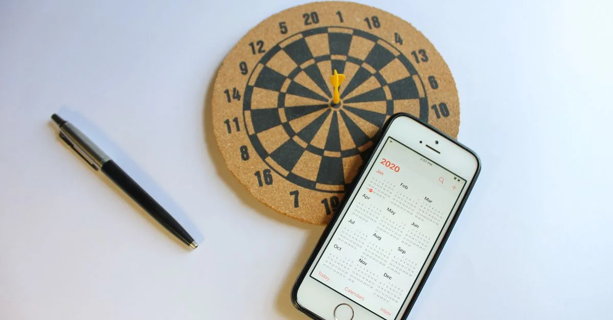 Un smartphone avec un calendrier 2020 à côté d'un tableau de fléchettes et d'un stylo sur une surface blanche.