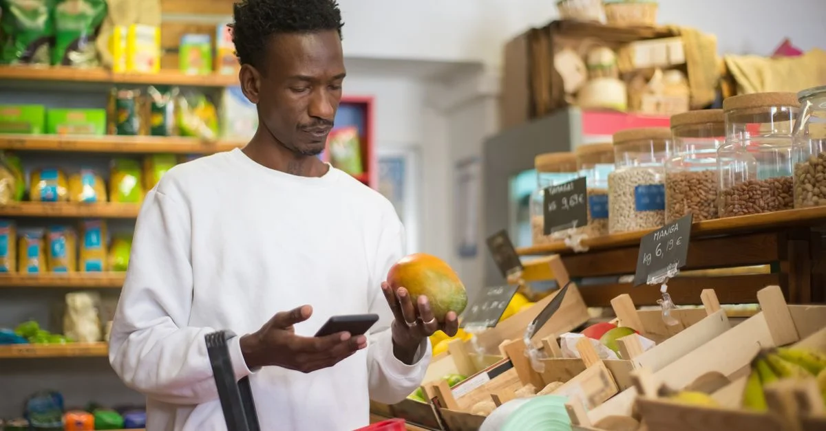 Un homme examine une mangue en faisant des courses avec un smartphone dans un supermarché.