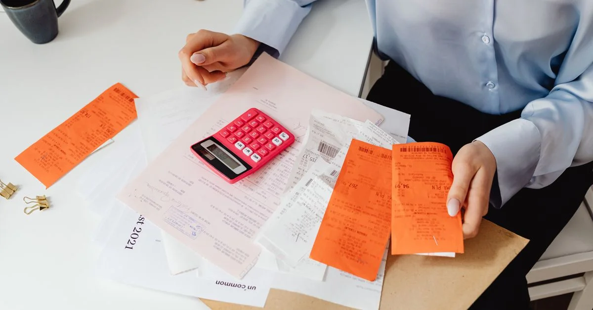 Une femme examine des reçus et calcule ses dépenses à un bureau avec une calculatrice rose.